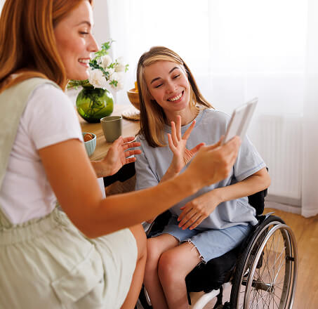 A woman in a wheelchair waving at a tablet device that another woman is holding