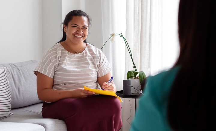 A woman sat on a sofa smiling to her practitioner who is facing away from camera